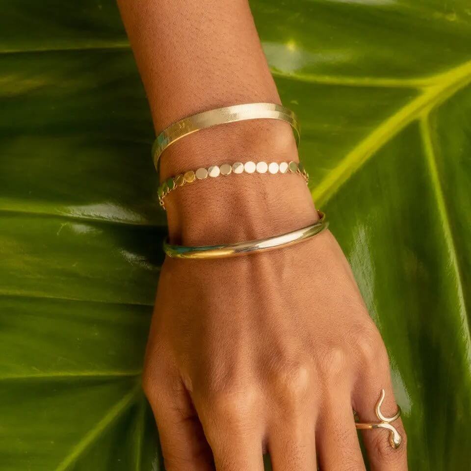 A close-up of a hand wearing a 10K gold bangle bracelet stacked with other bracelets, set against a green leaf backdrop.